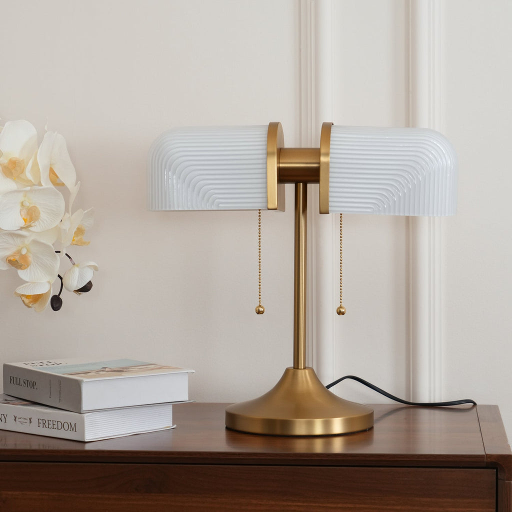 Close to the camera, this Clérachic gold table lamp with white ribbed shades stands on a walnut chest by white orchids.