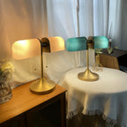 A lit Clérachic gold table lamp with white-glowing shades stands next to a second one with green shades, on a bedside table.