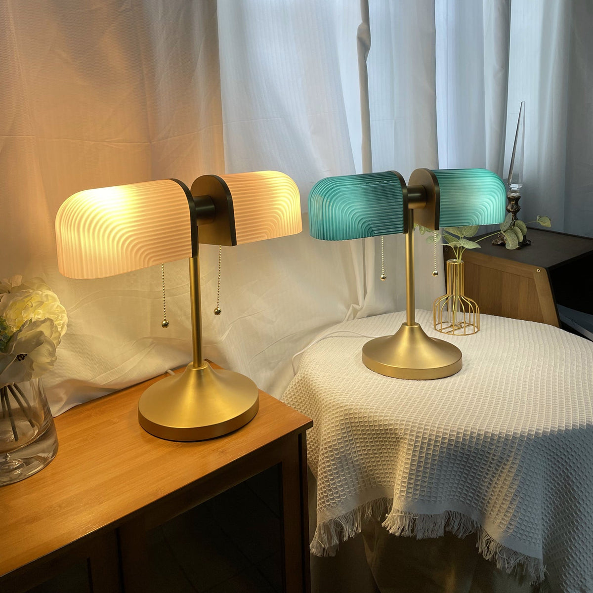 A lit Clérachic gold table lamp with white-glowing shades stands next to a second one with green shades, on a bedside table.