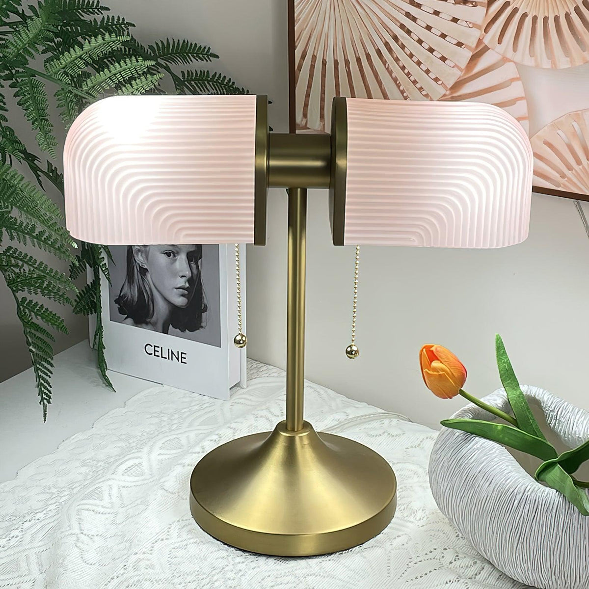 Close view of the pink ribbed glass shades and satin gold stem of this Clérachic table lamp, beside a fern and an orange tulip.