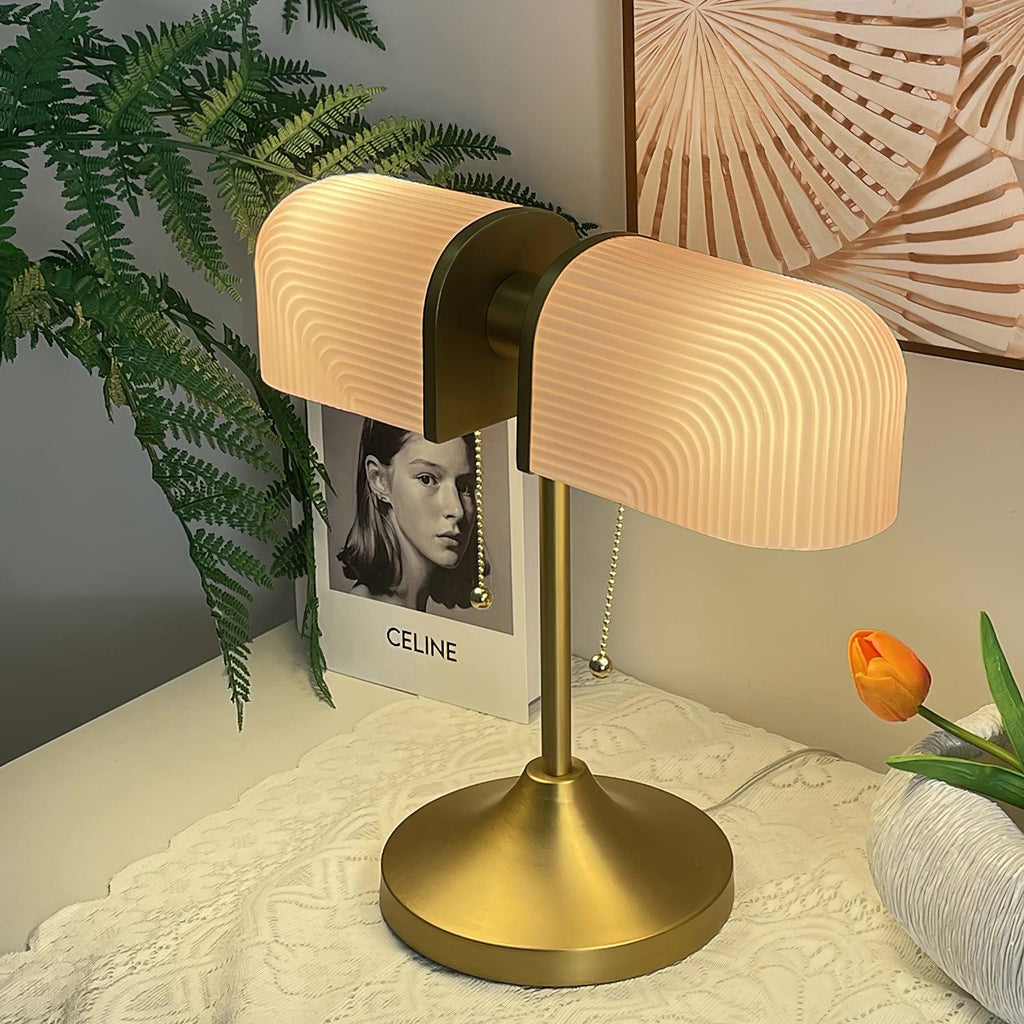 This Clérachic gold table lamp glows warm through its pink ribbed shades, with a fern, a book and an orange tulip behind.