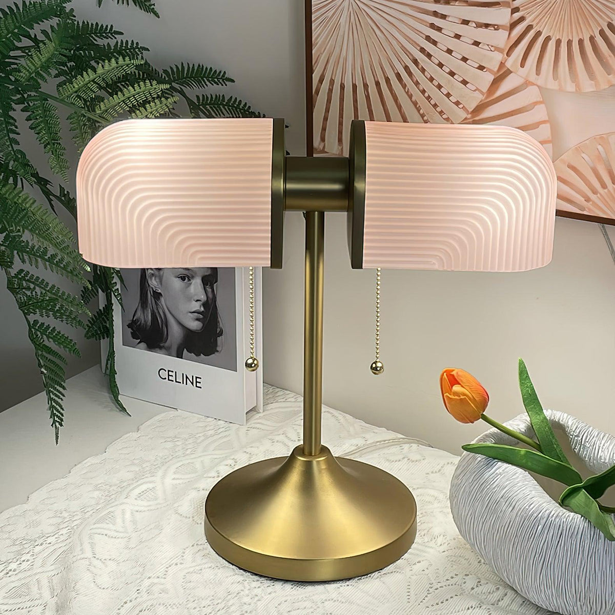 A Clérachic gold table lamp with pink ribbed shades is shown close up, one pull chain hanging down, against a patterned fan print.