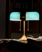 In a dark room, this Clérachic gold table lamp glows with bright blue-green shades that light a bookcase behind it.