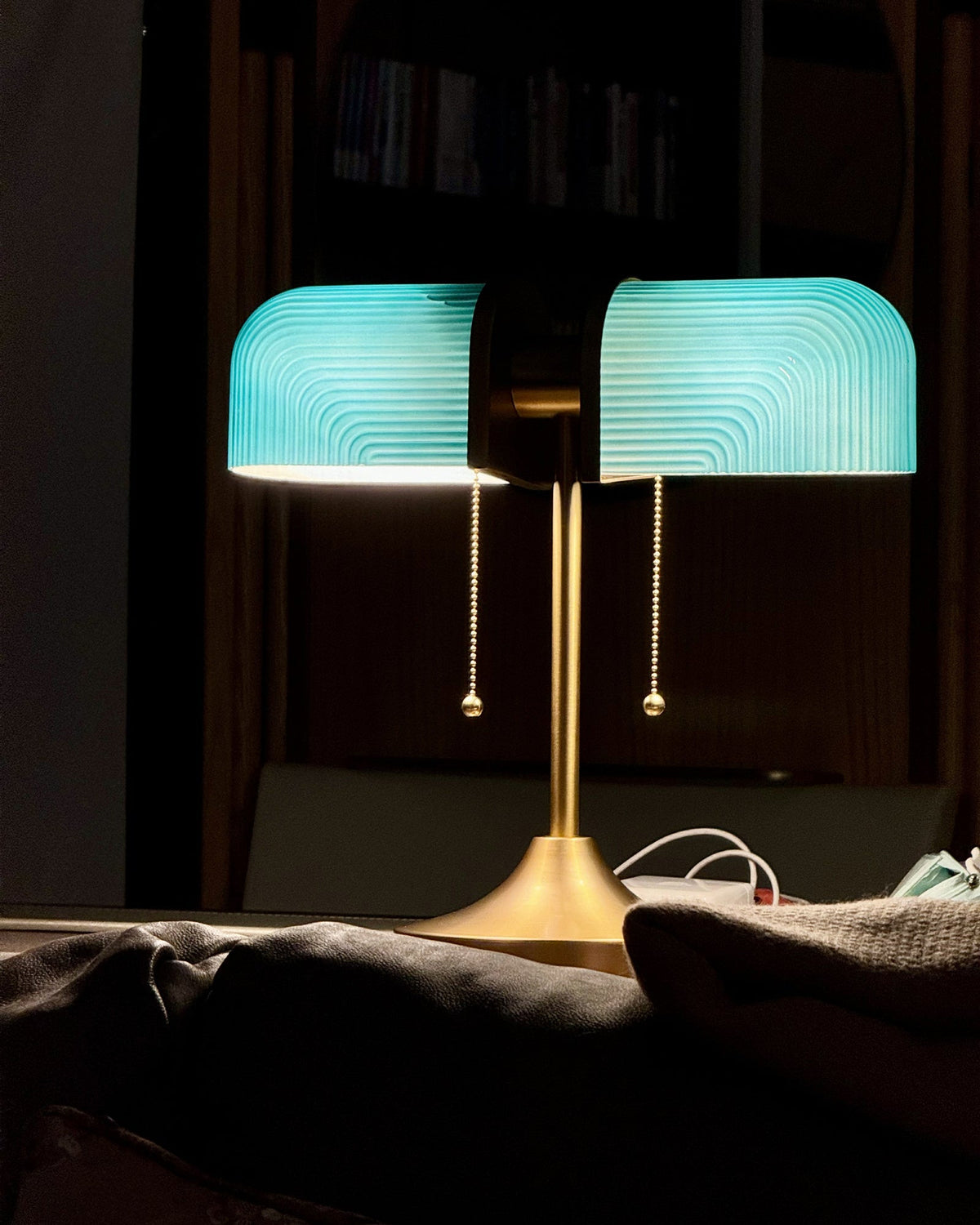 In a dark room, this Clérachic gold table lamp glows with bright blue-green shades that light a bookcase behind it.