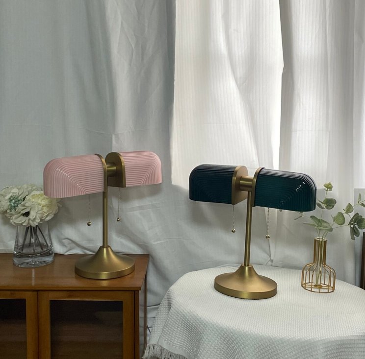 Two Clérachic gold table lamps, one with pink shades and one with dark green, stand on a wooden cabinet and a draped table.