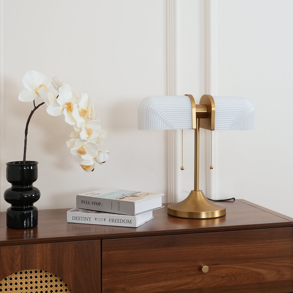 Shown here, a Clérachic gold table lamp with white ribbed shades sits at the right of a walnut chest, with a black glass vase and white orchids.