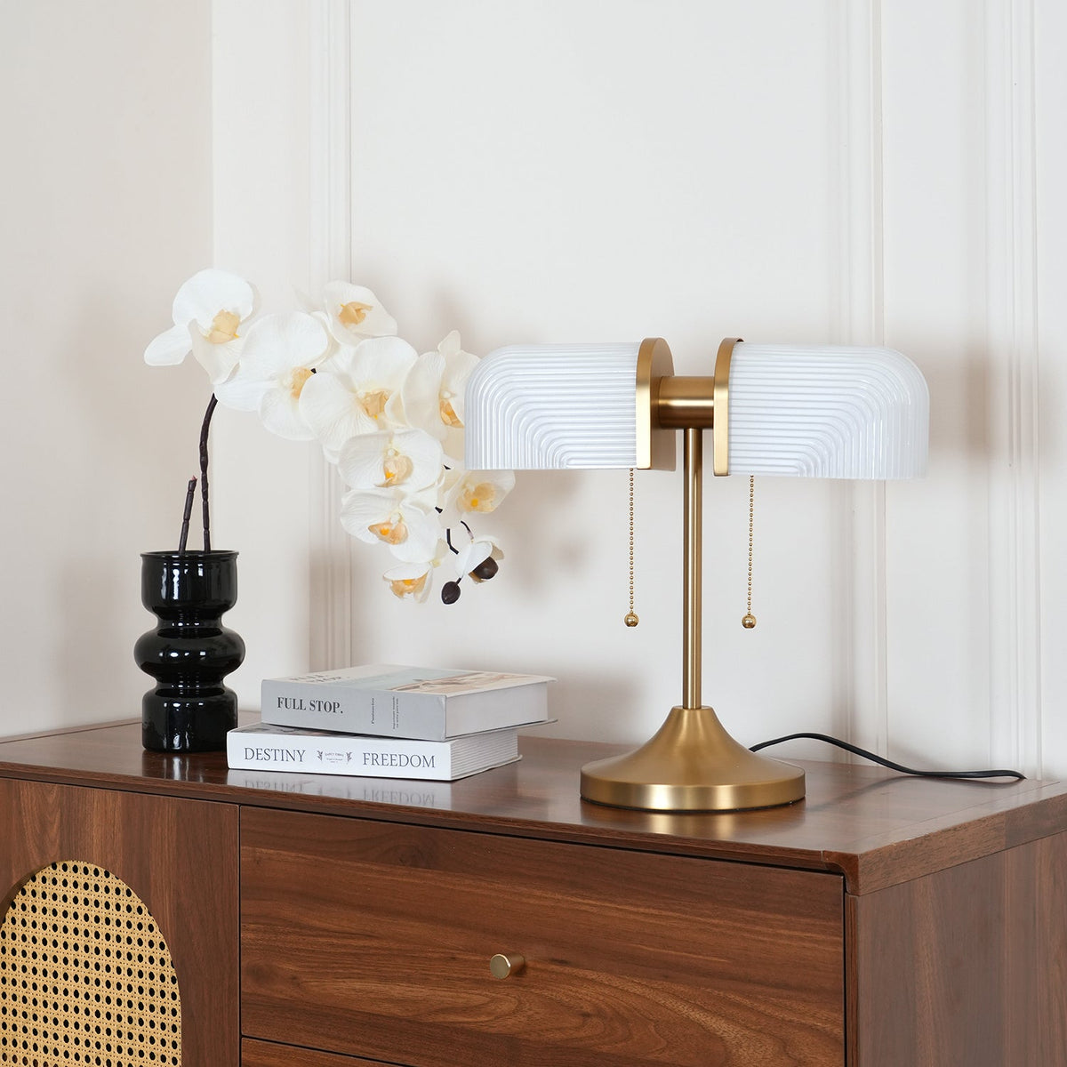 This Clérachic gold table lamp with white ribbed shades stands on a walnut chest with a rattan panel, below a spray of orchids.