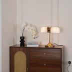 A wider view of this Clérachic gold table lamp lit warm, on a walnut cabinet with a rattan arch door and white orchids.