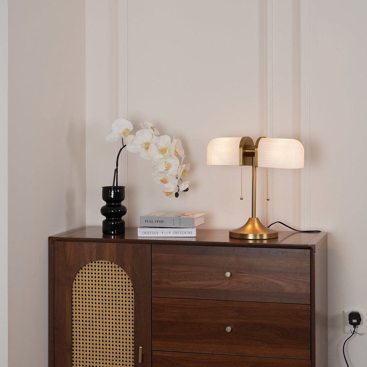 A wider view of this Clérachic gold table lamp lit warm, on a walnut cabinet with a rattan arch door and white orchids.
