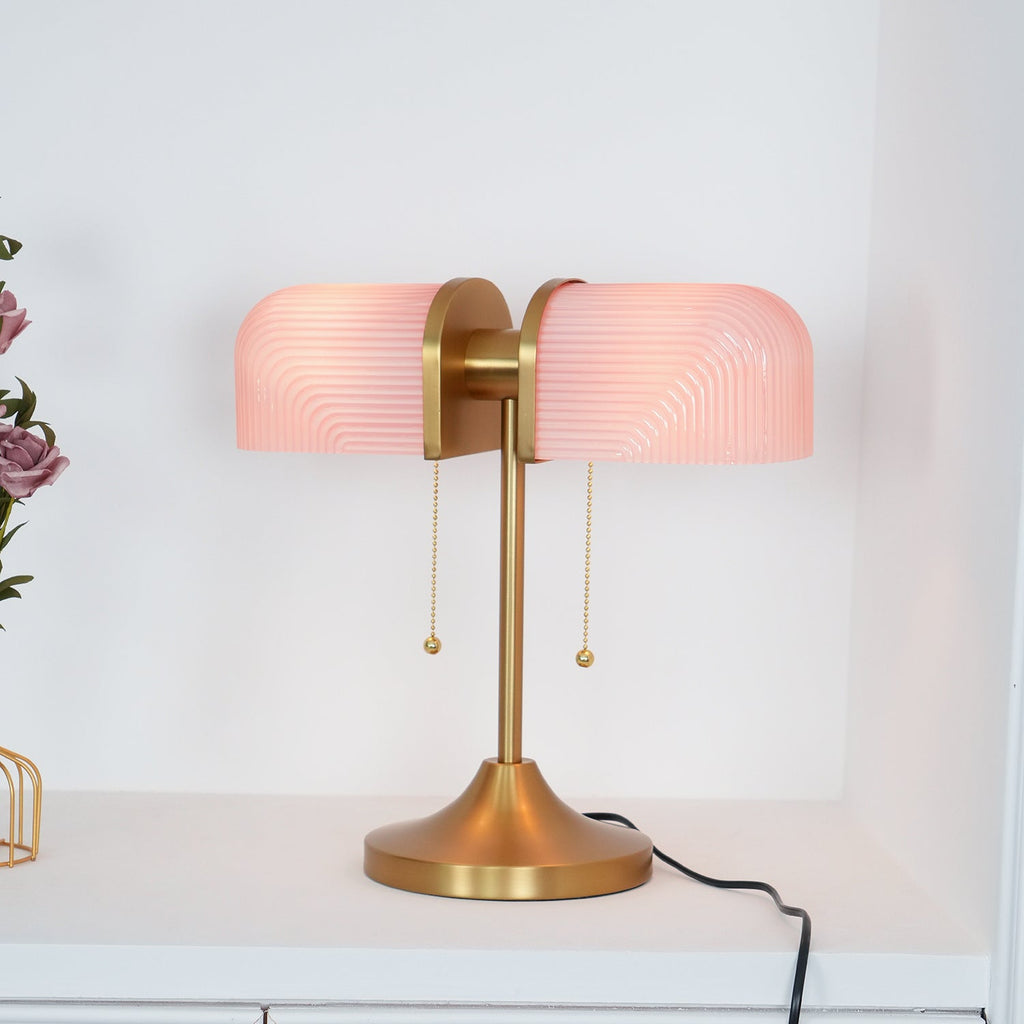 This Clérachic gold table lamp with pink ribbed shades glows on a white shelf, its black cord trailing to the right.