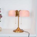 This Clérachic gold table lamp with pink ribbed shades glows on a white shelf, its black cord trailing to the right.