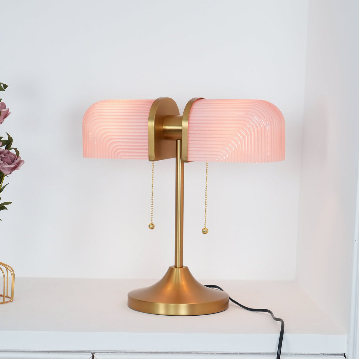 This Clérachic gold table lamp with pink ribbed shades glows on a white shelf, its black cord trailing to the right.