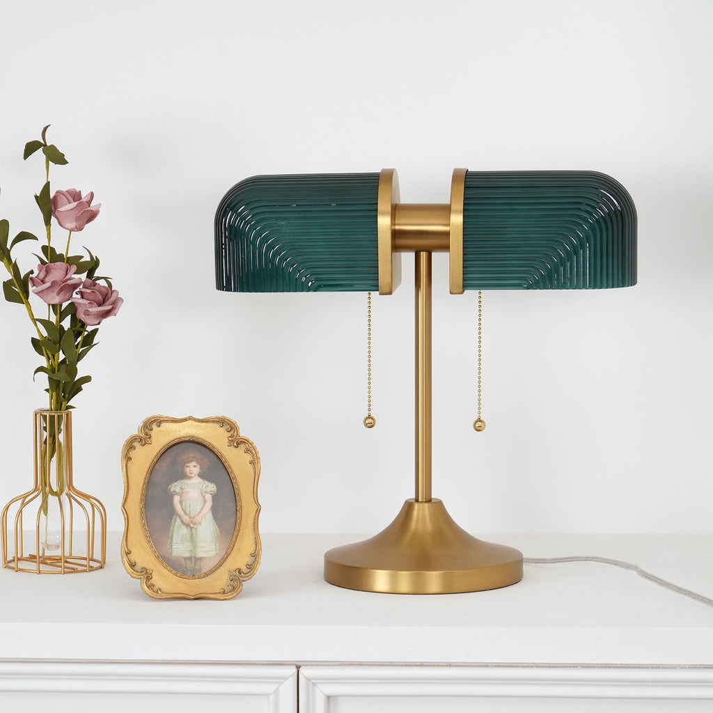 A Clérachic gold table lamp with deep green ribbed glass shades beside a small gilt-framed portrait and a vase of pink roses.