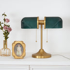 A Clérachic gold table lamp with deep green ribbed glass shades beside a small gilt-framed portrait and a vase of pink roses.