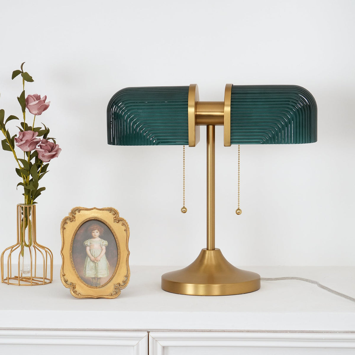 A Clérachic gold table lamp with deep green ribbed glass shades beside a small gilt-framed portrait and a vase of pink roses.