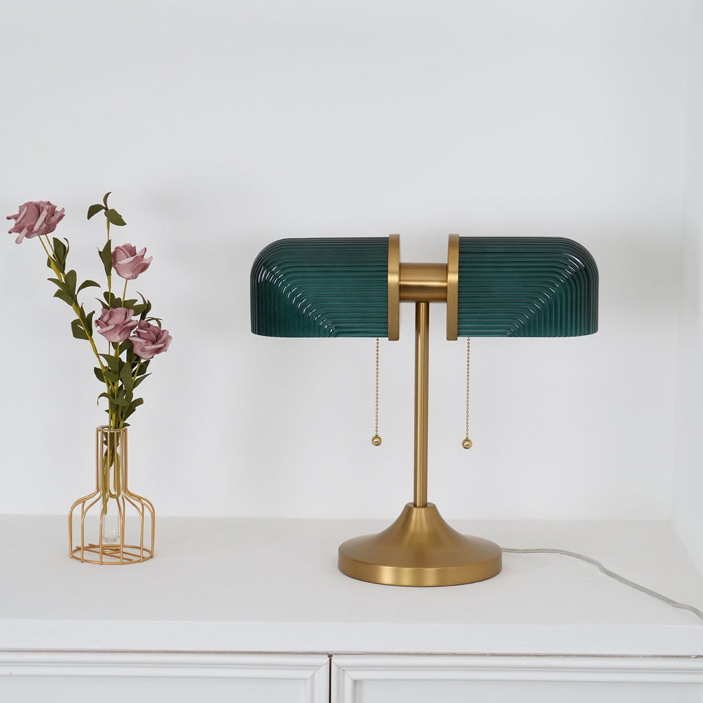 Pictured here, a Clérachic gold table lamp with dark green ribbed shades stands on a white cabinet, beside a wire vase of mauve roses.