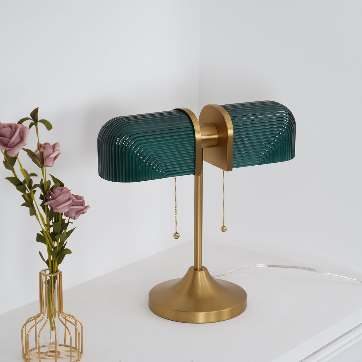 Angled view of this Clérachic gold table lamp with dark green shades and a flared satin gold base, beside a vase of roses.