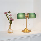 This Clérachic gold table lamp lights its green ribbed shades from within, on a white cabinet beside a vase of pink roses.
