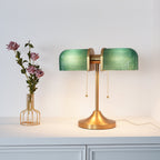 A lit Clérachic gold table lamp with green ribbed shades stands to the right of a wire vase of pink roses, on a white cabinet.