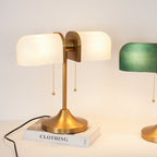 Shown here, a Clérachic gold table lamp with glowing white ribbed shades sits on a book, with a green-shaded lamp partly in view at the right.