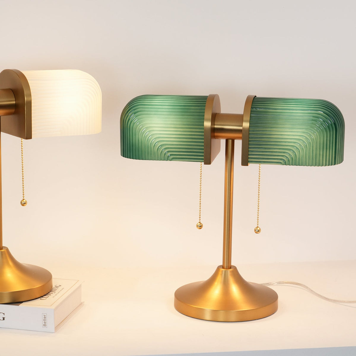 A green-shaded Clérachic gold table lamp stands beside a white-shaded one partly in view, both lit on a white cabinet.