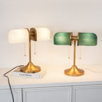 Two lit Clérachic gold table lamps, one with white shades and one with green, stand side by side on a white cabinet.