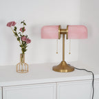 In this photo, a Clérachic gold table lamp with pink ribbed shades, unlit, stands beside a wire vase of roses on a white cabinet.