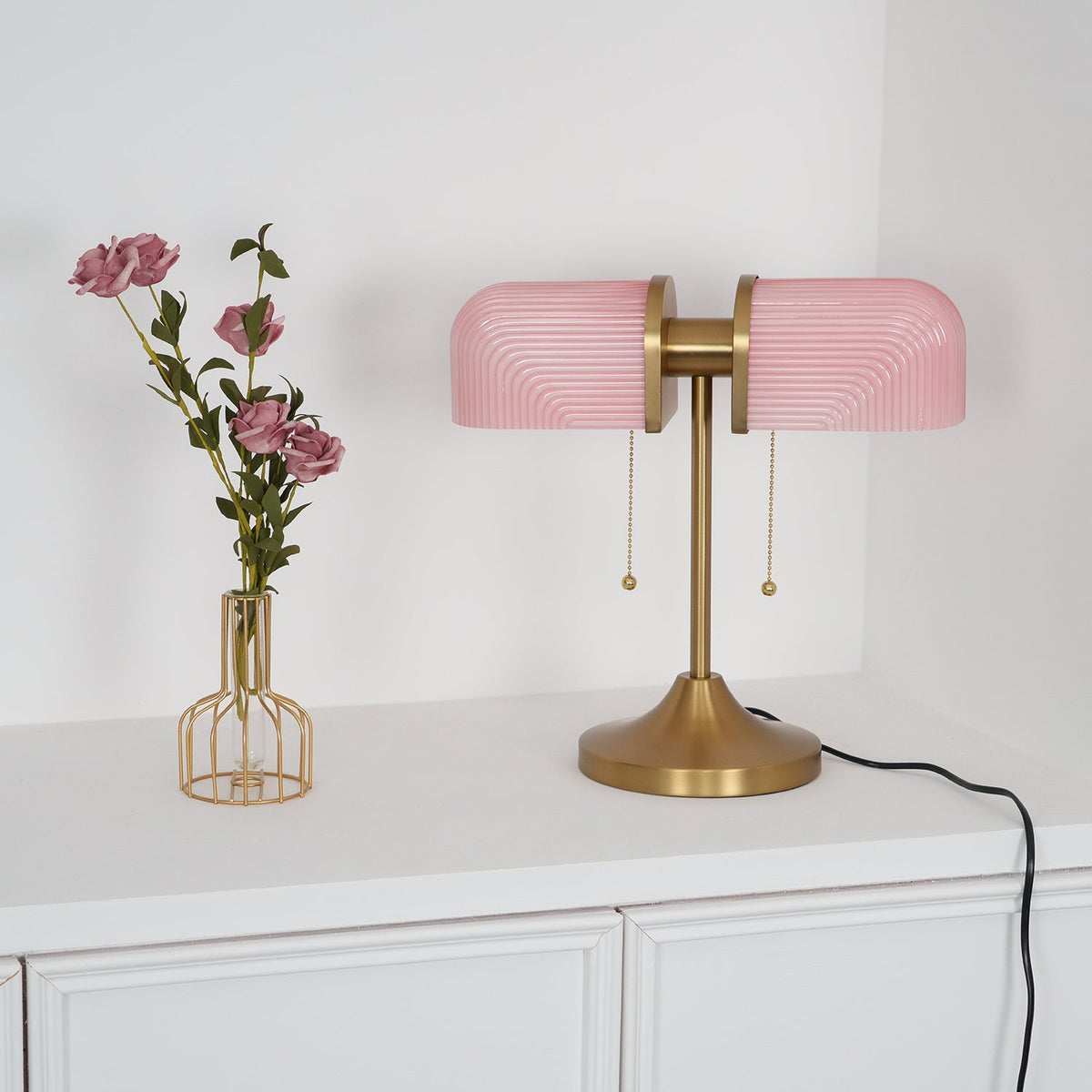 In this photo, a Clérachic gold table lamp with pink ribbed shades, unlit, stands beside a wire vase of roses on a white cabinet.