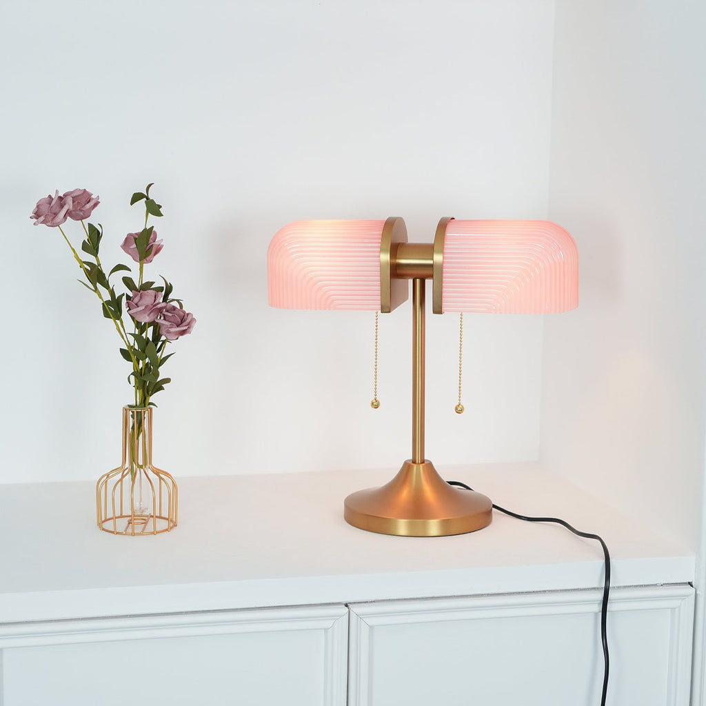 Soft light comes through the pink ribbed shades of this Clérachic gold table lamp, with a vase of roses at its left.