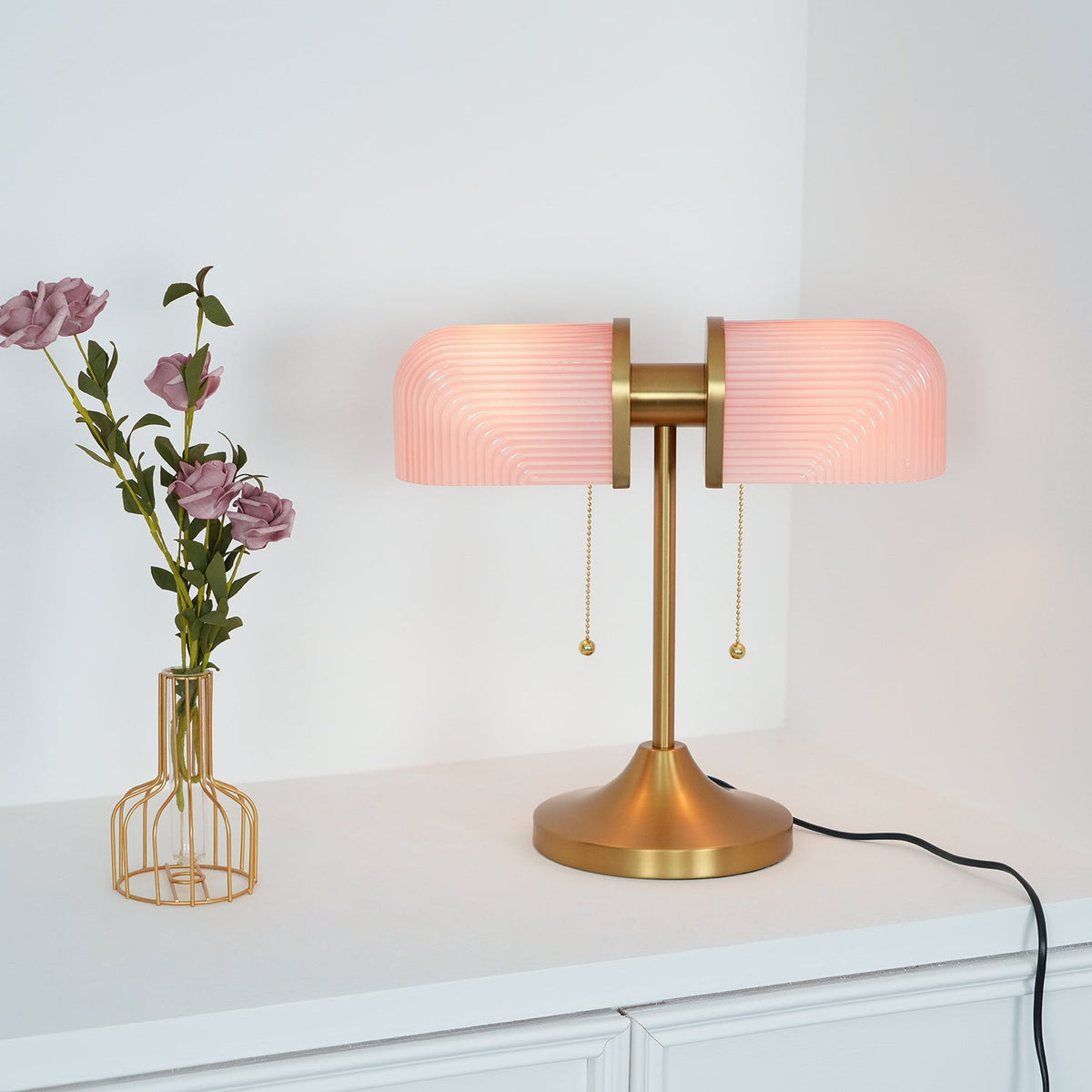 A closer view of this Clérachic gold table lamp with pink shades glowing, a vase of roses and a black cord beside it.