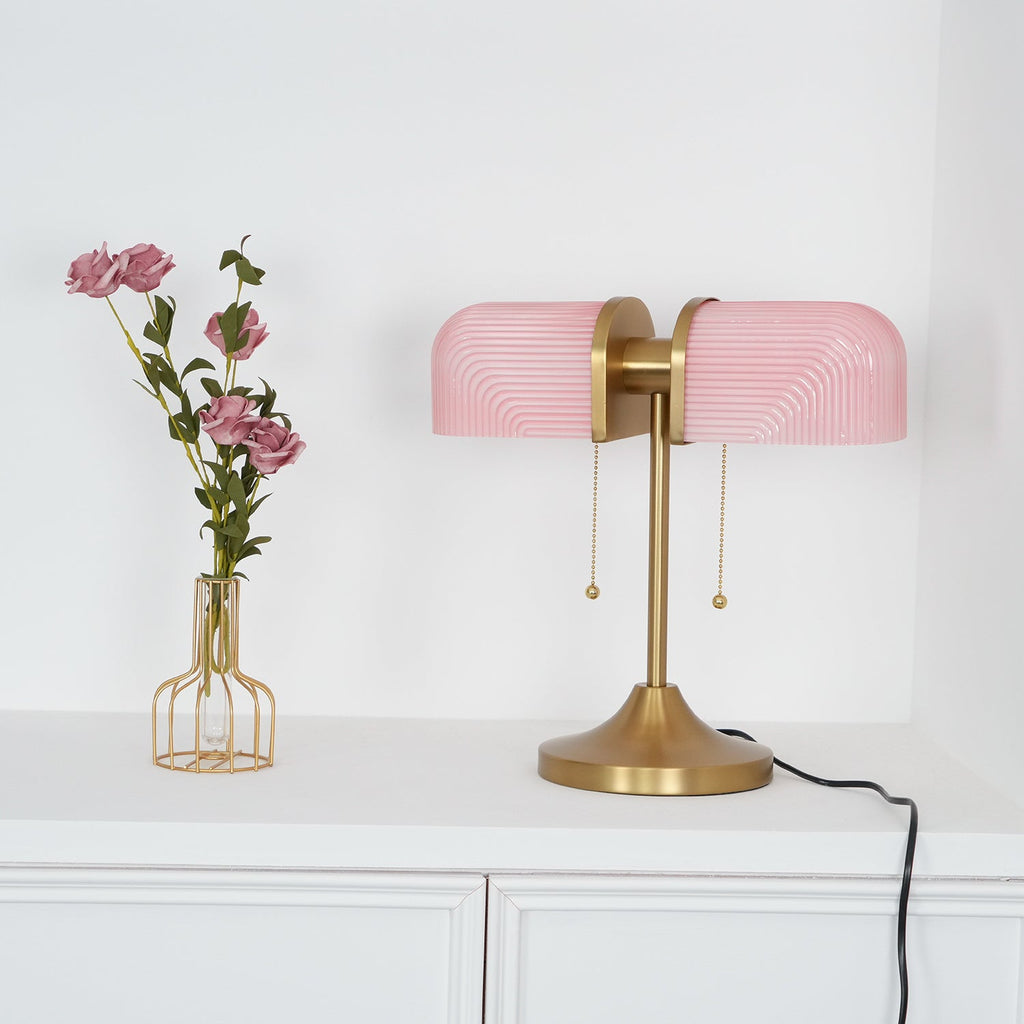 A Clérachic gold table lamp with pink ribbed shades stands on a white cabinet, its black cord curling down at the right.