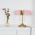 A Clérachic gold table lamp with pink ribbed shades stands on a white cabinet, its black cord curling down at the right.