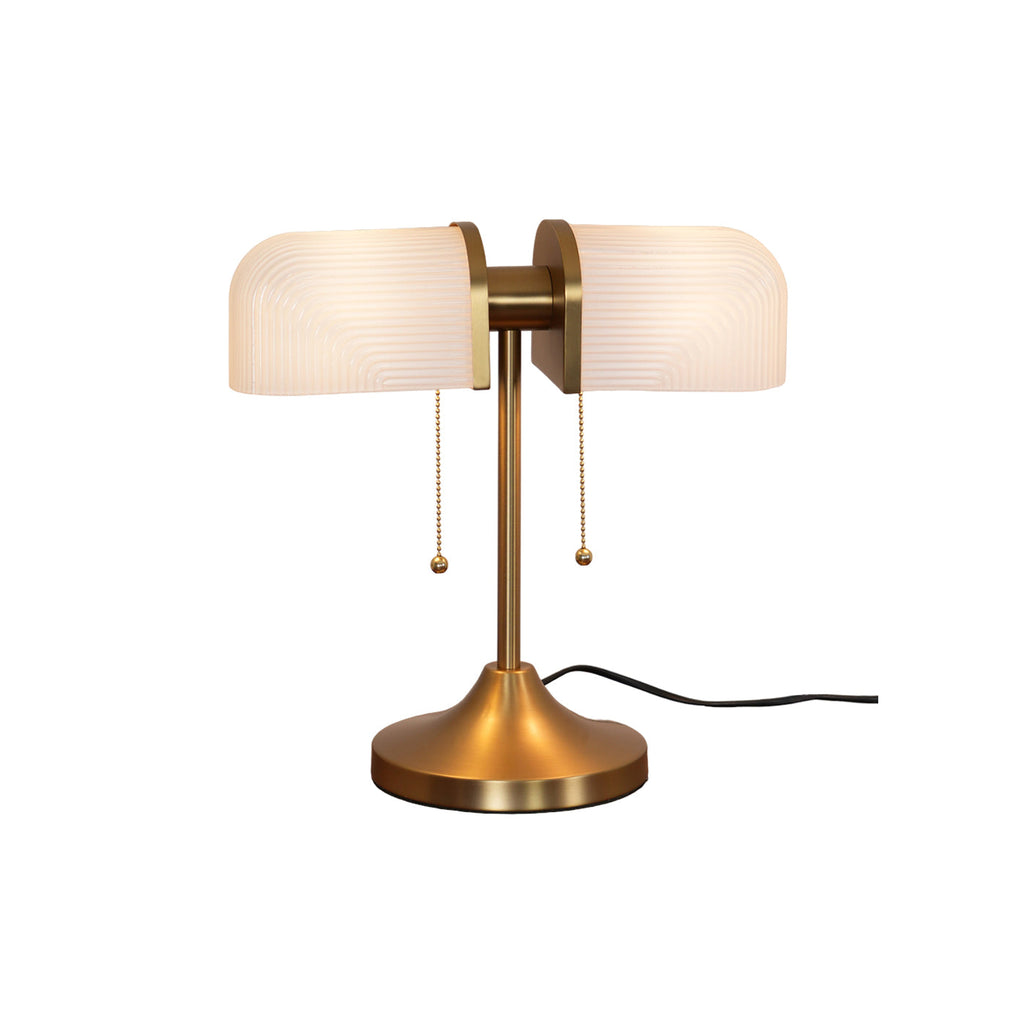 Pictured here, a Clérachic gold table lamp with warm white ribbed shades, lit, with a black cord trailing from its flared gold base.