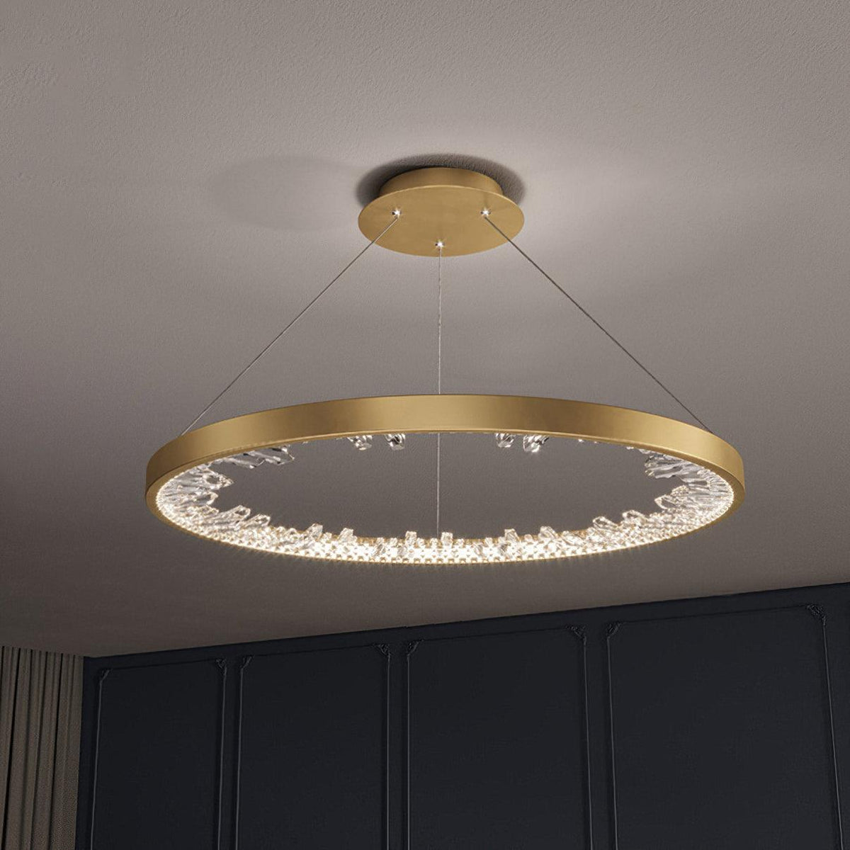 Gold Clérachic ring pendant light with a round ceiling plate and crossed cables, hanging in front of a dark panelled wall.