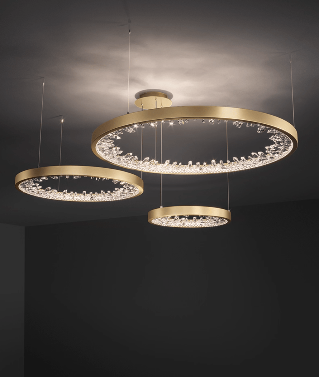 Three gold Clérachic ring pendants of different sizes lit at different heights in a dark room, their clear glass pieces sparkling.