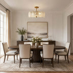 A Clérachic ceiling light with gold fittings hangs above a dark oval dining table with cream chairs on a patterned rug.