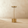 Clérachic cordless table lamp in warm brushed gold, disc shade on a slim stem above a cylindrical base against a plaster wall.