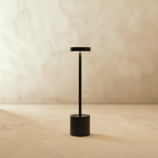 Clérachic cordless table lamp in matte black, disc shade and slim stem set on a round base against a plaster wall.