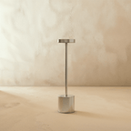 Clérachic cordless table lamp in brushed silver, disc shade on a slim stem above a cylindrical base against a plaster wall.