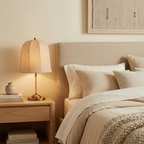 A lit Clérachic linen table lamp on a pale oak bedside table, beside a bed with cream bedding and a chunky knit throw.