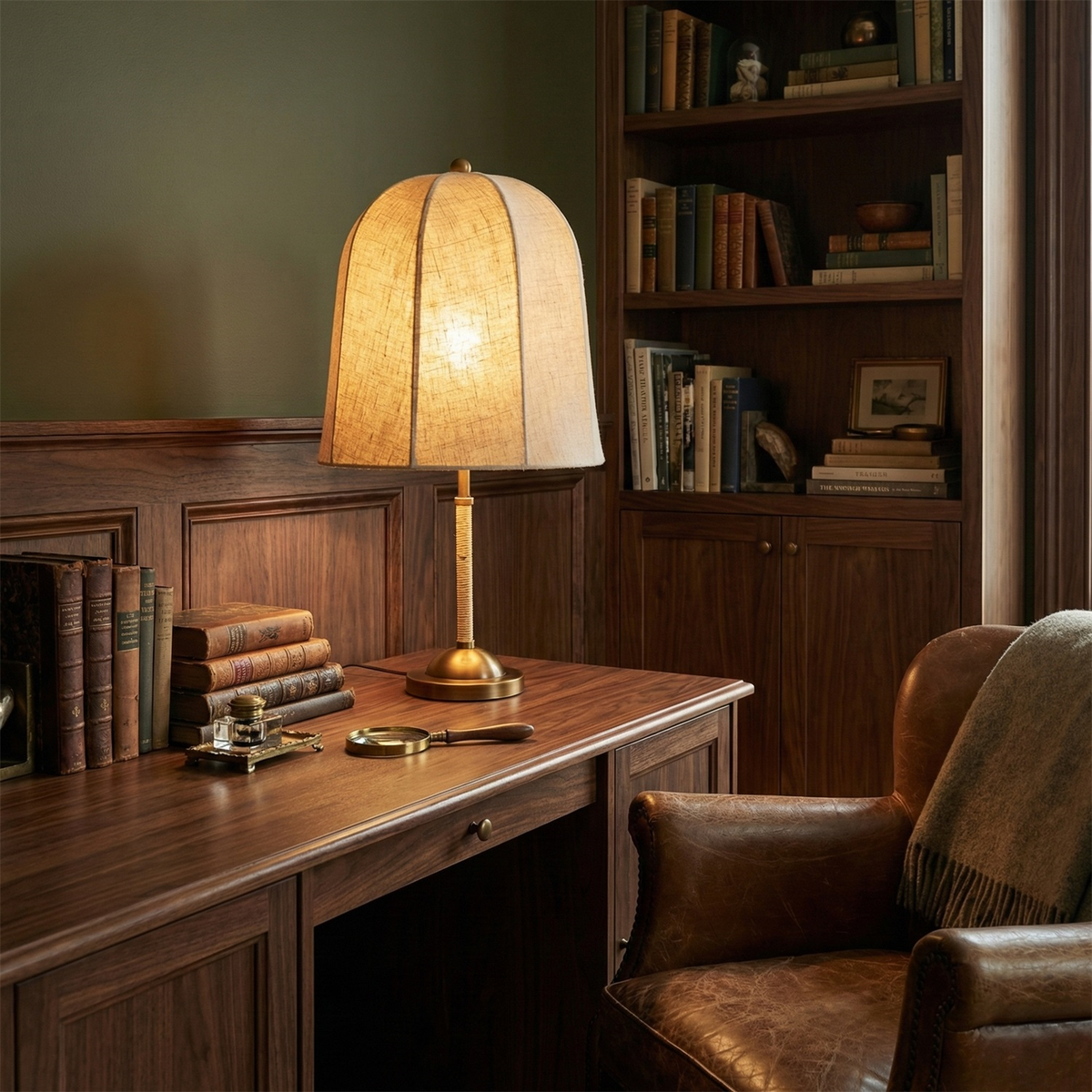 A Clérachic linen table lamp glows on a dark walnut desk, with leather-bound books, a brown leather armchair and green walls.