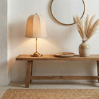 A Clérachic linen table lamp glows on a rustic wooden bench, with a round mirror, dried pampas grass in a vase and a jute rug.