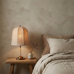 A lit Clérachic linen table lamp on a rustic wooden stool beside a bed with linen bedding, with a small vase of dried stems.