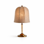 A Clérachic table lamp with a ribbed linen shade lit from within, on a slim stem and gold base, on a white backdrop.