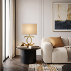 A Clérachic gold table lamp with a cream rectangular shade on a round black side table, beside a boucle sofa and an abstract print.