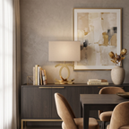 Clérachic gold table lamp with a cream shade on a dark wooden sideboard, beside a stack of books, a stone vase and a dining table.