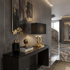 A Clérachic gold table lamp with a black shade on a dark marble console, beside a black vase, books and a brass globe, in a marble-floored hall.