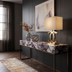 Clérachic gold table lamp with a cream shade on a purple-veined marble console, with a vase of dried stems and a brass sculpture.