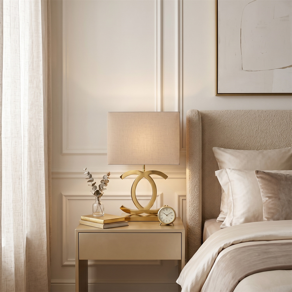 Lit Clérachic gold table lamp with a cream shade on a pale bedside table beside a bed with cream bedding and a small clock.