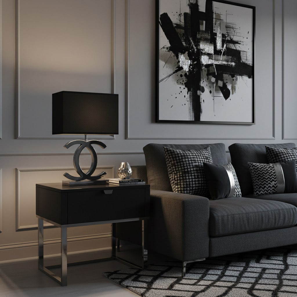 A Clérachic table lamp with a black shade and a dark metal base on a black bedside table, next to a dark grey sofa and a black and white print.